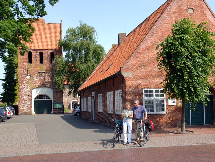 07_056.JPG Ein altes Backsteingebäude mit roten Dachziegeln und Bäumen davor. Zwei Radfahrer stehen lachend daneben.