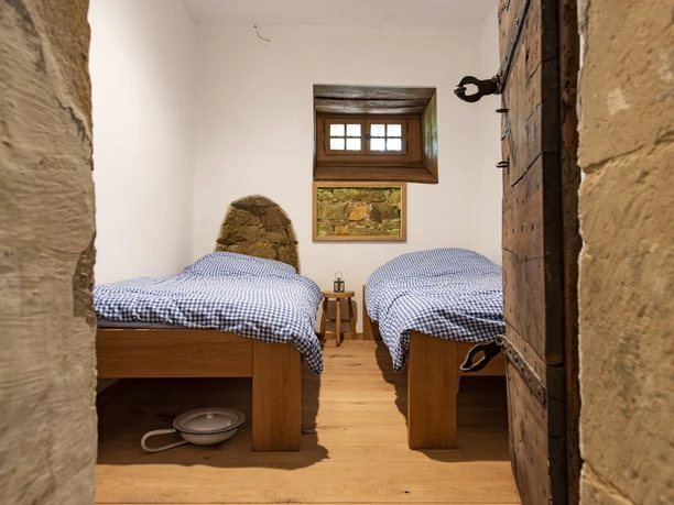 amtsgefaengnis-SGFuerstenau-Osnabruecker-Land-Erlebnis-Ausflug-Uebernachtung Zwei Einzelbetten in rustikalem Raum mit Holzböden und kleinem Fenster oberhalb.