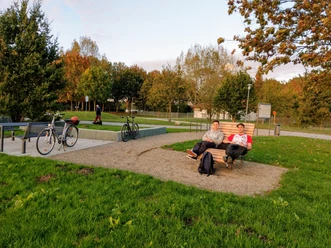 Zwei Personen entspannen auf einer Liegebank an einem Rastplatz mit Fahrrädern auf grünem Gras.