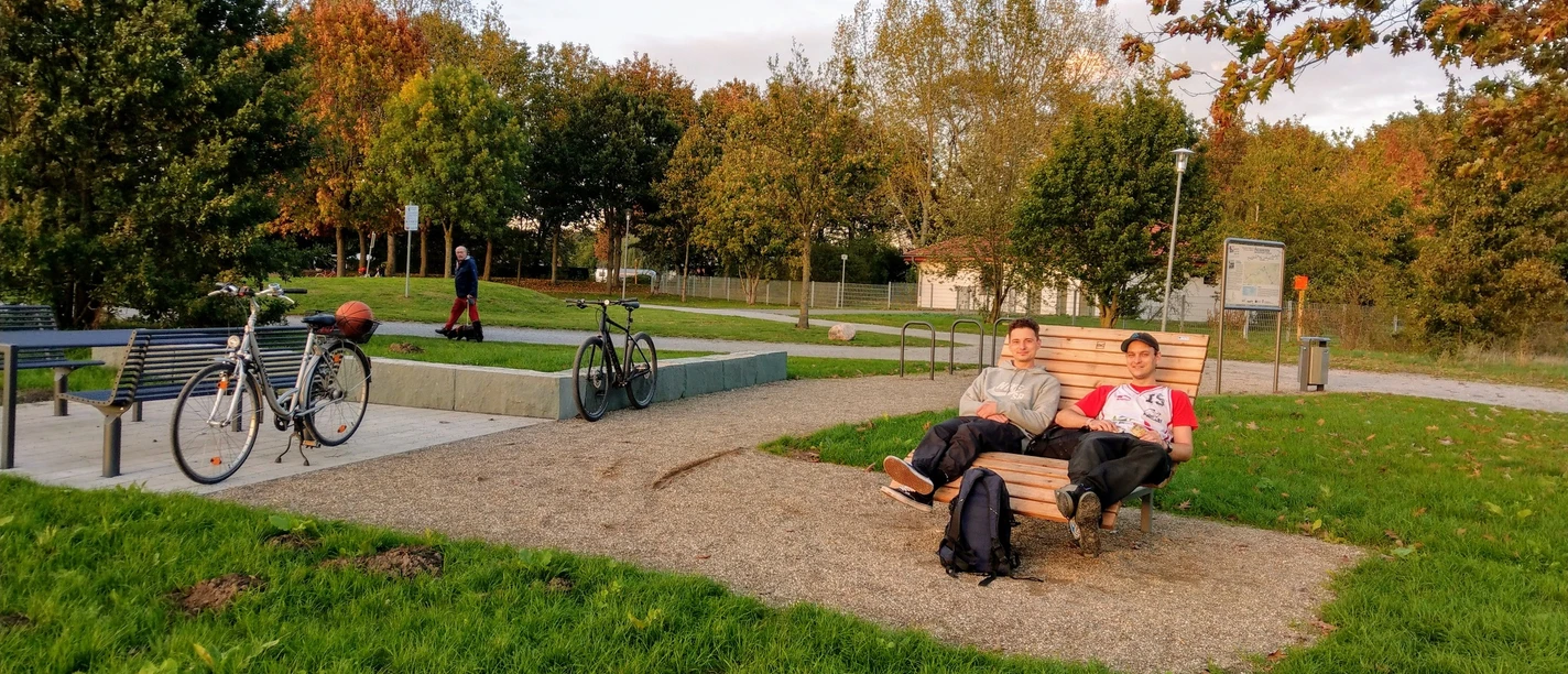 Zwei Personen entspannen auf einer Liegebank an einem Rastplatz mit Fahrrädern auf grünem Gras.