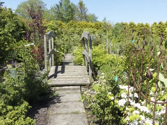 Gartencenter Schittenhelm der Baumschule Leezdorf Holzbrücke inmitten eines blühenden Gartens mit farbenreicher Bepflanzung und üppigem Grün.