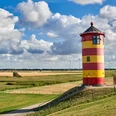 Pilsumer Leuchtturm Roter und gelber Leuchtturm auf grünem Deich vor weitem Himmel mit Wolken und Windrädern im Hintergrund.