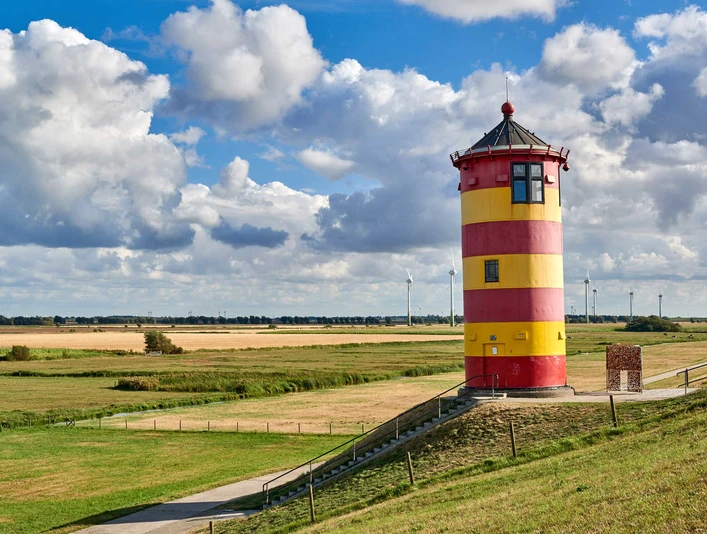 Pilsumer Leuchtturm Roter und gelber Leuchtturm auf grünem Deich vor weitem Himmel mit Wolken und Windrädern im Hintergrund.