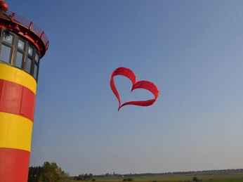 Zweites Eheversprechen im Pilsumer Leuchtturm Rot-gelber Leuchtturm vor blauem Himmel mit herzförmigem roten Drachen über grüner Landschaft.