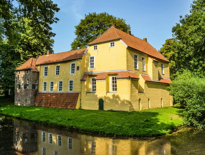 Manningaburg Pewsum Gelb verputzte Backsteinburg mit rotem Ziegeldach, umgeben von Wassergraben und alten Bäumen.