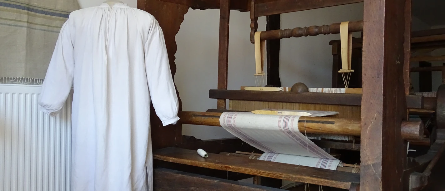 Ein historischer Webstuhl mit einem weißen, nahtlosen Hemd daneben, zeigt traditionelle Webkunst.