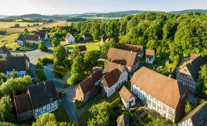 Detmold-2023-023-LWL Freilichtmuseum-Teutoburger-Wald-Tourismus-D-Ketz-CC-BY-SA.jpg Luftaufnahme zeigt ein Freilichtmuseum in Detmold mit Fachwerkhäusern und grüner, hügeliger Landschaft.