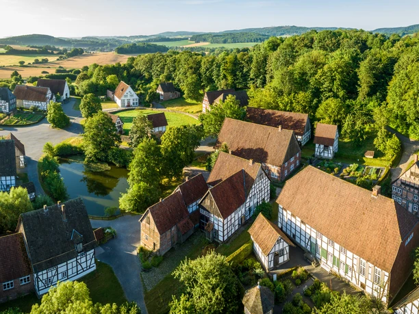 Detmold-2023-023-LWL Freilichtmuseum-Teutoburger-Wald-Tourismus-D-Ketz-CC-BY-SA.jpg Luftaufnahme zeigt ein Freilichtmuseum in Detmold mit Fachwerkhäusern und grüner, hügeliger Landschaft.