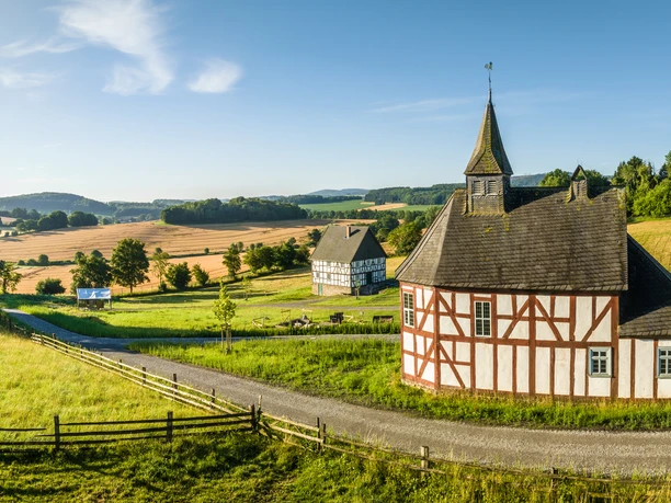 Detmold-2023-033-LWL Freilichtmuseum-Teutoburger-Wald-Tourismus-D-Ketz.jpg Fachwerk-Kapelle mit Turm in grüner Landschaft bei Detmold, umgeben von Feldern und Hügeln.