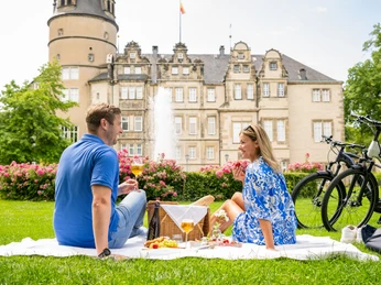 Ein Paar genießt ein Picknick auf einer Wiese vor einem historischen Schloss in Detmold.