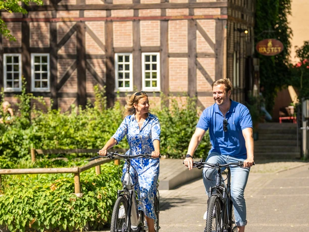 Detmold-2023-177-Lippisches Landesmuseum-Teutoburger-Wald-Tourismus-D-Ketz.jpg Zwei Personen radeln entspannt auf einem gepflasterten Weg, vorbei an einem Fachwerkgebäude.