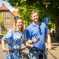Paar lächelt beim Fahrradfahren vor Fachwerkhäusern in sonniger Straßenszene in Detmold.
