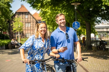 Detmold-2023-181-Lippisches Landesmuseum-Teutoburger-Wald-Tourismus-D-Ketz.jpg Paar lächelt beim Fahrradfahren vor Fachwerkhäusern in sonniger Straßenszene in Detmold.