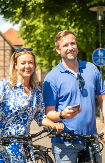 Detmold-2023-181-Lippisches Landesmuseum-Teutoburger-Wald-Tourismus-D-Ketz.jpg Paar lächelt beim Fahrradfahren vor Fachwerkhäusern in sonniger Straßenszene in Detmold.