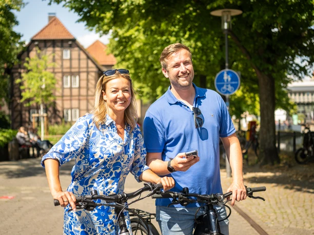 Detmold-2023-181-Lippisches Landesmuseum-Teutoburger-Wald-Tourismus-D-Ketz.jpg Paar lächelt beim Fahrradfahren vor Fachwerkhäusern in sonniger Straßenszene in Detmold.