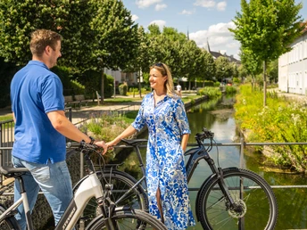 Detmold-2023-186-Am Wallgraben-Teutoburger-Wald-Tourismus-D-Ketz.jpg Ein Mann und eine Frau stehen mit Fahrrädern lächelnd auf einer Brücke neben einem Fluss.