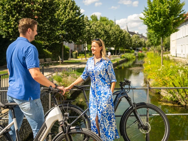 Detmold-2023-186-Am Wallgraben-Teutoburger-Wald-Tourismus-D-Ketz.jpg Ein Mann und eine Frau stehen mit Fahrrädern lächelnd auf einer Brücke neben einem Fluss.