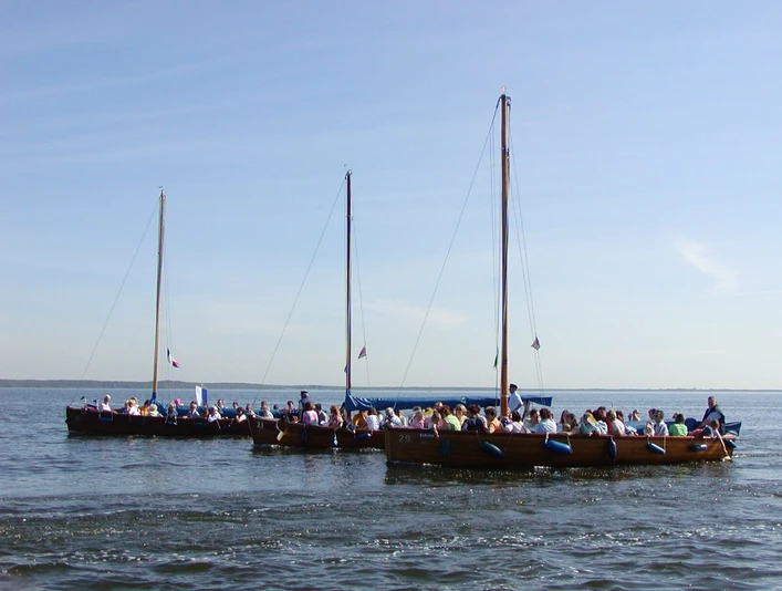 Drei Auswanderer Drei Boote voll besetzt mit Menschen fahren bei klarem Himmel über ein weites Gewässer.Three boats full of people sail across a wide stretch of water under a clear sky.Tre både fulde af mennesker sejler over et stort vandområde under en klar himmel.Drie boten vol mensen varen over een breed stuk water onder een heldere hemel.