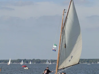 Auswanderer unter Segel Ein historisches Holzsegelboot gleitet sanft über das ruhige Wasser, während Menschen an Bord zusehen.A historic wooden sailing boat glides gently over the calm water while people watch on board.En historisk sejlbåd af træ glider blidt hen over det rolige vand, mens folk ser på om bord.Een historische houten zeilboot glijdt zachtjes over het kalme water terwijl mensen aan boord toekijken.