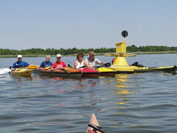 Kajaks Postboje Fünf Kajakfahrer auf einem ruhigen See paddeln gemeinsam vor einer gelben Postboje unter blauem Himmel.