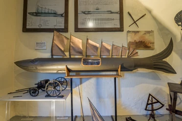 Modell eines Fisches mit Segeln, umgeben von Zeichnungen und nautischen Instrumenten an der Wand.