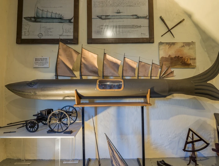 Modell eines Fisches mit Segeln, umgeben von Zeichnungen und nautischen Instrumenten an der Wand.