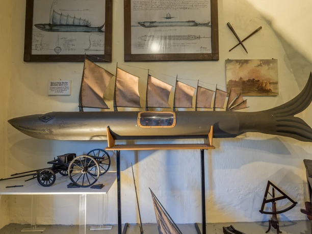 Modell "Steinhuder Hecht" Modell eines Fisches mit Segeln, umgeben von Zeichnungen und nautischen Instrumenten an der Wand.