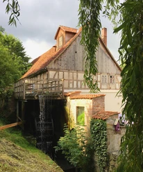 Historische Wassermühle mit sichtbarem Mühlrad und Fachwerkhaus, umgeben von üppigem Grün.