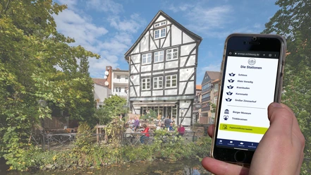 Wasserwege-Zimmerhof Handydisplay zeigt Stationsübersicht, Fachwerkhaus mit Terrasse am Wasser