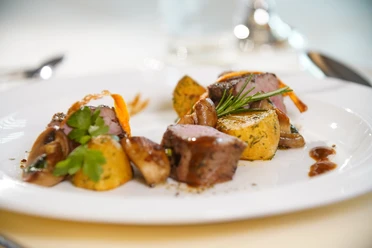 Gastro Beispielbild TOL .jpg Köstlich angerichtetes Fleischgericht mit karamellisierten Kartoffeln, Pilzen und Blättchen auf weißem Teller.