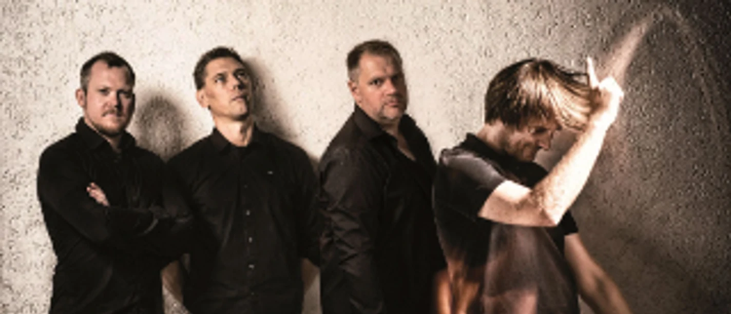 q-bic-live-funkband-im-filou.png Vierköpfige Funkband, alle in dunklen Hemden, posieren vor einer grauen Wand, einer tanzt freudig.