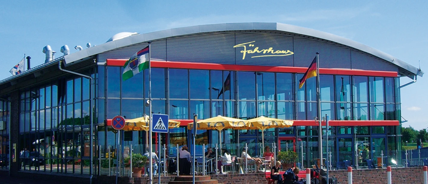 f-hrhause-terrasse-mit-logo Das Fährhaus am Emder Außenhafen: Modernes Gebäude mit bunten Sonnenschirmen.