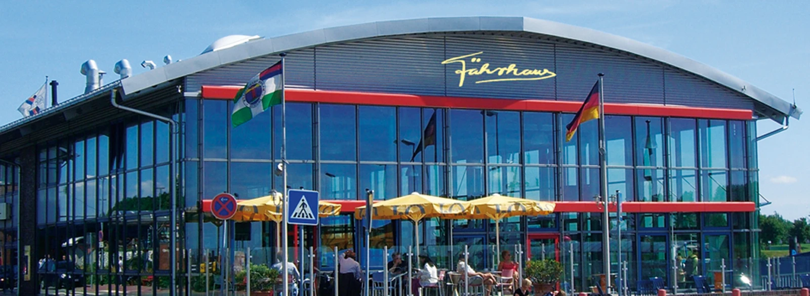 f-hrhause-terrasse-mit-logo Das Fährhaus am Emder Außenhafen: Modernes Gebäude mit bunten Sonnenschirmen.