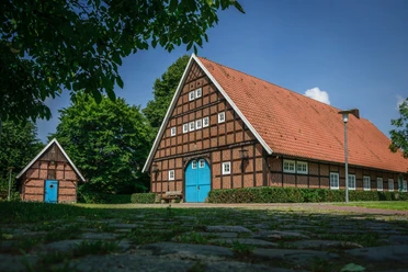 Heimathof Sögel Historisches Fachwerkhaus mit rotem Ziegeldach und blauer Torfront, umgeben von grünen Bäumen.
