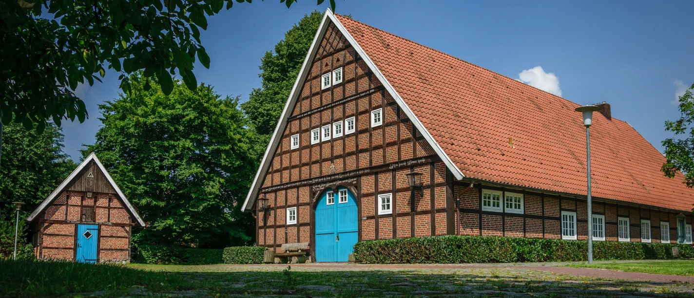 Heimathof Sögel Historisches Fachwerkhaus mit rotem Ziegeldach und blauer Torfront, umgeben von grünen Bäumen.