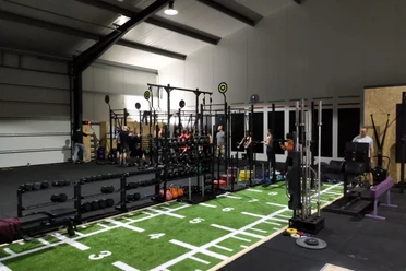 Crossfit Geräte Innenraum einer Halle mit Crossfit-Geräten, darunter Langhanteln und Rudergeräte; Personen trainieren.