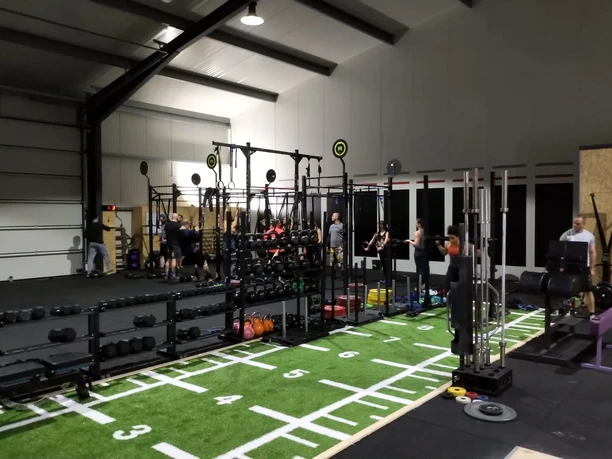 Crossfit Geräte Innenraum einer Halle mit Crossfit-Geräten, darunter Langhanteln und Rudergeräte; Personen trainieren.