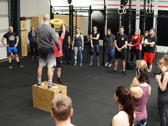 CrossFit Gruppe Eine CrossFit-Gruppe steht in einem Fitnessstudio im Kreis. Vorne auf einem Holzkasten zeigt ein Trainer stolz eine Gewichtsscheibe.