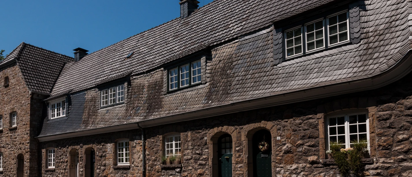 Das Bild zeigt eine historische Reihenhausfassade aus Stein mit Schieferdach und weißen Fenstern.