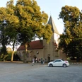 Apostelkirche Ovenstädt - Petershagen Apostelkirche Ovenstädt umgeben von hohen Bäumen, Parkplatz mit weißem Auto davor, Besuchende sichtbar.