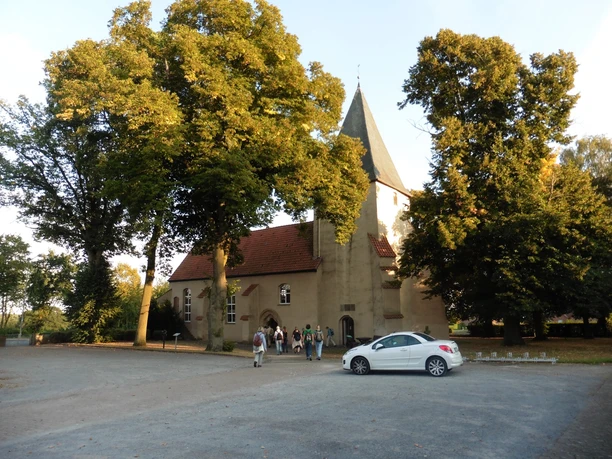 Apostelkirche Ovenstädt - Petershagen Apostelkirche Ovenstädt umgeben von hohen Bäumen, Parkplatz mit weißem Auto davor, Besuchende sichtbar.