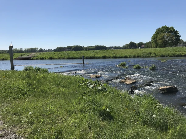 Sohlengleite Quakenbrück Fluss mit grünem Ufer, Steine im Wasser, blauer Himmel, ruhige Natur, horizontale Perspektive.