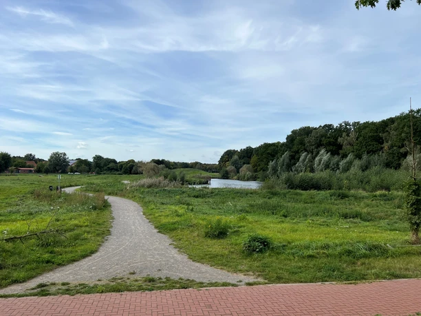 Ein Wanderweg aus Ziegelsteinen führt durch eine grüne Wiesenlandschaft zu einem See, Wald im Hintergrund.