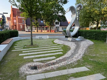 Moderne Skulptur in urbaner Umgebung mit gepflastertem Weg und parkähnlicher Grünanlage.