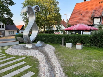 Wave Metallene Skulptur einer geschwungenen Welle in einem kleinen Park, flankiert von Bäumen und Häusern.