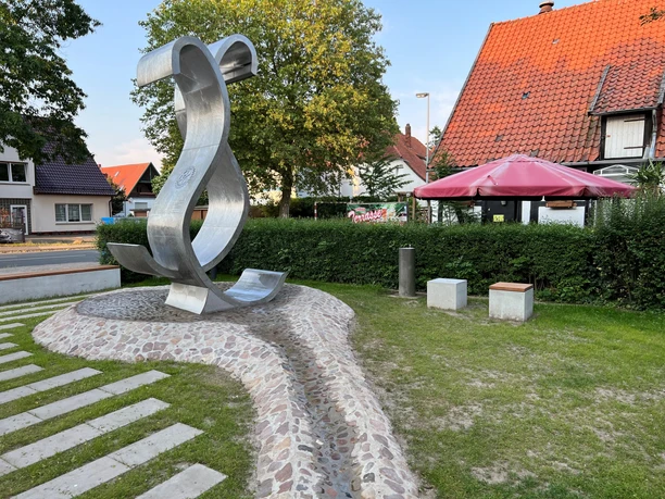 Wave Metallene Skulptur einer geschwungenen Welle in einem kleinen Park, flankiert von Bäumen und Häusern.