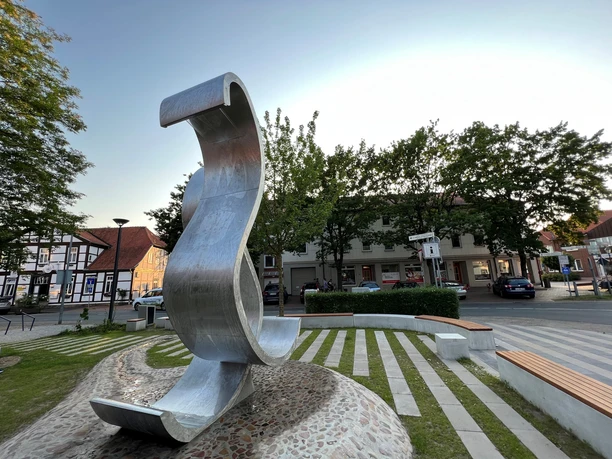 Moderne Skulptur einer geschwungenen Metallwelle auf einem Platz mit Bäumen und umliegenden Gebäuden.