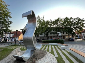 Wave Moderne Skulptur einer geschwungenen Metallwelle auf einem Platz mit Bäumen und umliegenden Gebäuden.