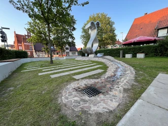 Moderne Metallskulptur in einem grünen Parkbereich mit Bäumen und umliegenden historischen Gebäuden.