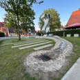 Wave Moderne Metallskulptur in einem grünen Parkbereich mit Bäumen und umliegenden historischen Gebäuden.